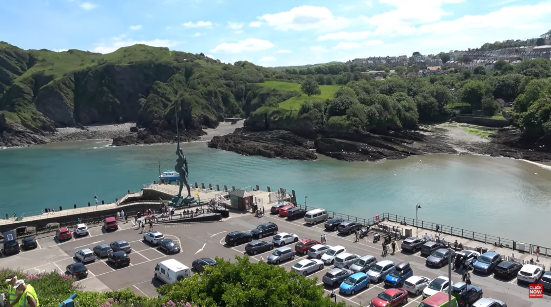 parking with cars, a statue of a woman, river, and hills with trees