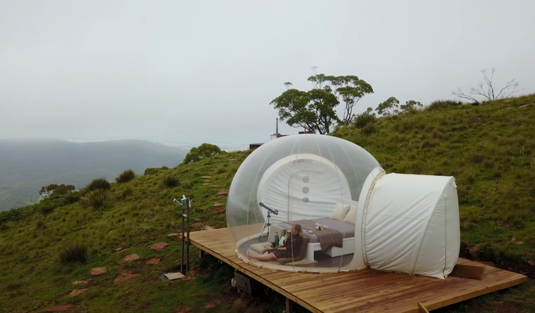 a bed and woman inside the transparent bubble hotel among the hills