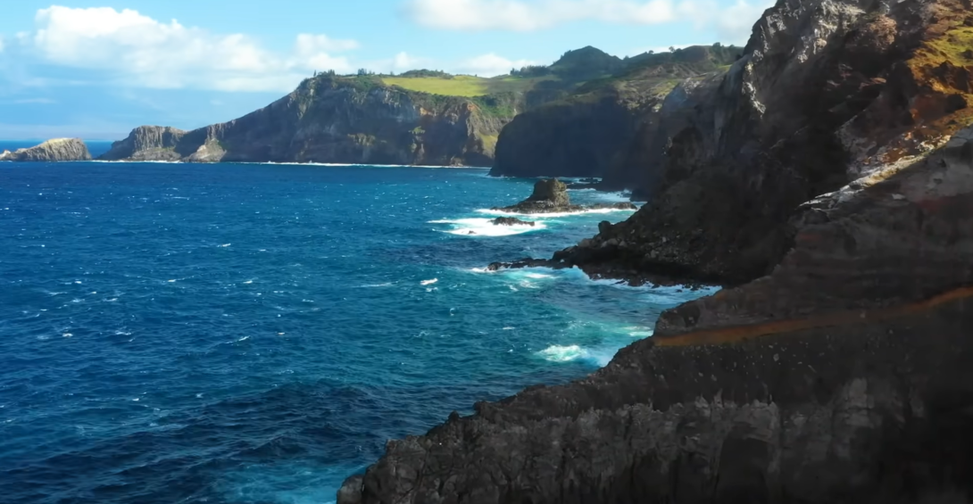 ocean with waves and mountains with trees on it, clouds above