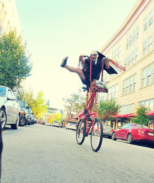 a crazy guy in glasses wearing a nun’s outfit riding an ugly-looking red bike