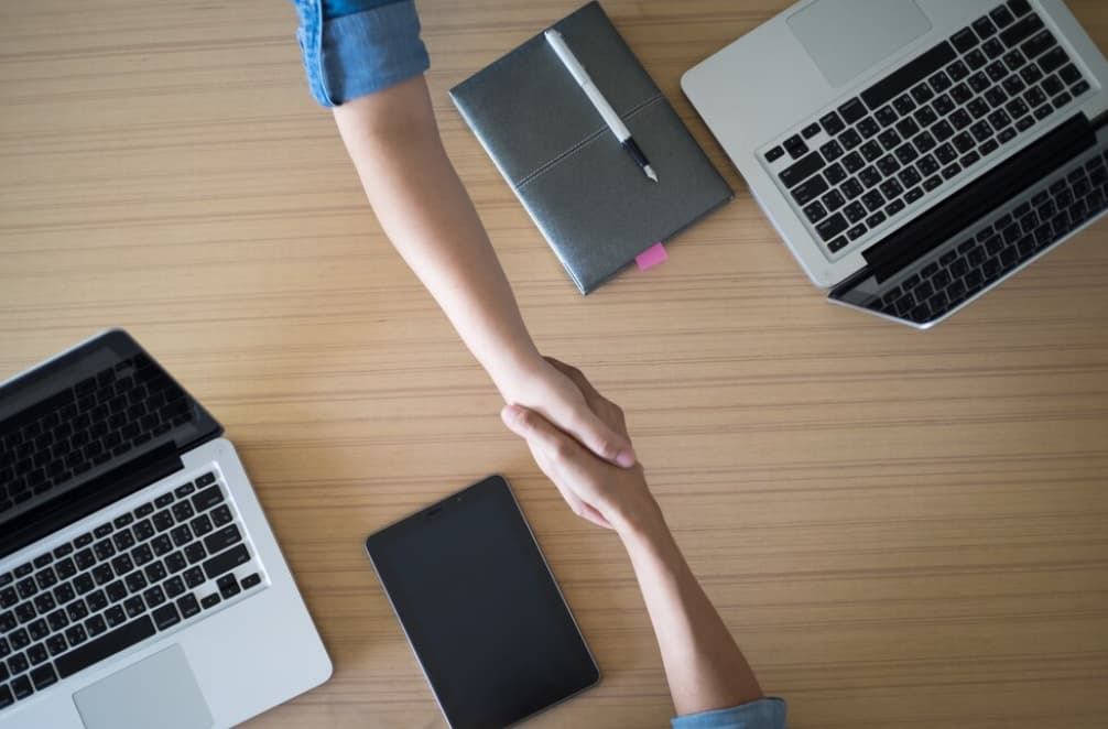 two hands holding each other in front of the table with two opened laptop and notebook on it