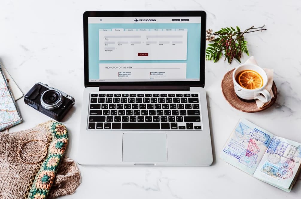 A laptop with a travel booking website, camera, and a cozy knit bag on a white surface